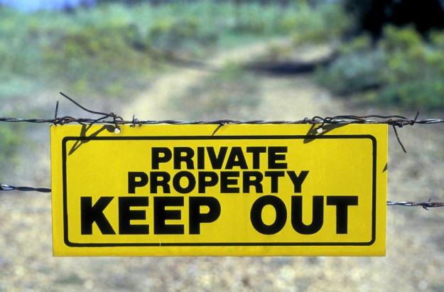 A yellow and black "Private Property Keep Out" sign attached to a barbed wire gate stretched across a dirt road.